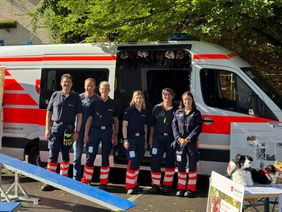Auf dem Foto sieht man mehrere Personen des Besuchs- und Begleitungsdienstes mit Herz und Hund zusammen mit Ihren Hunden. Im Hintergrund ist ein Einsatzfahrzeug der Malteser zu erkennen. Im Vordergrund sind Übungsgeräte für die Rettungshunde, darunter eine Wippe zu erkennen sowie ein Tisch auf dem Plüschhunde zu sehen sind.