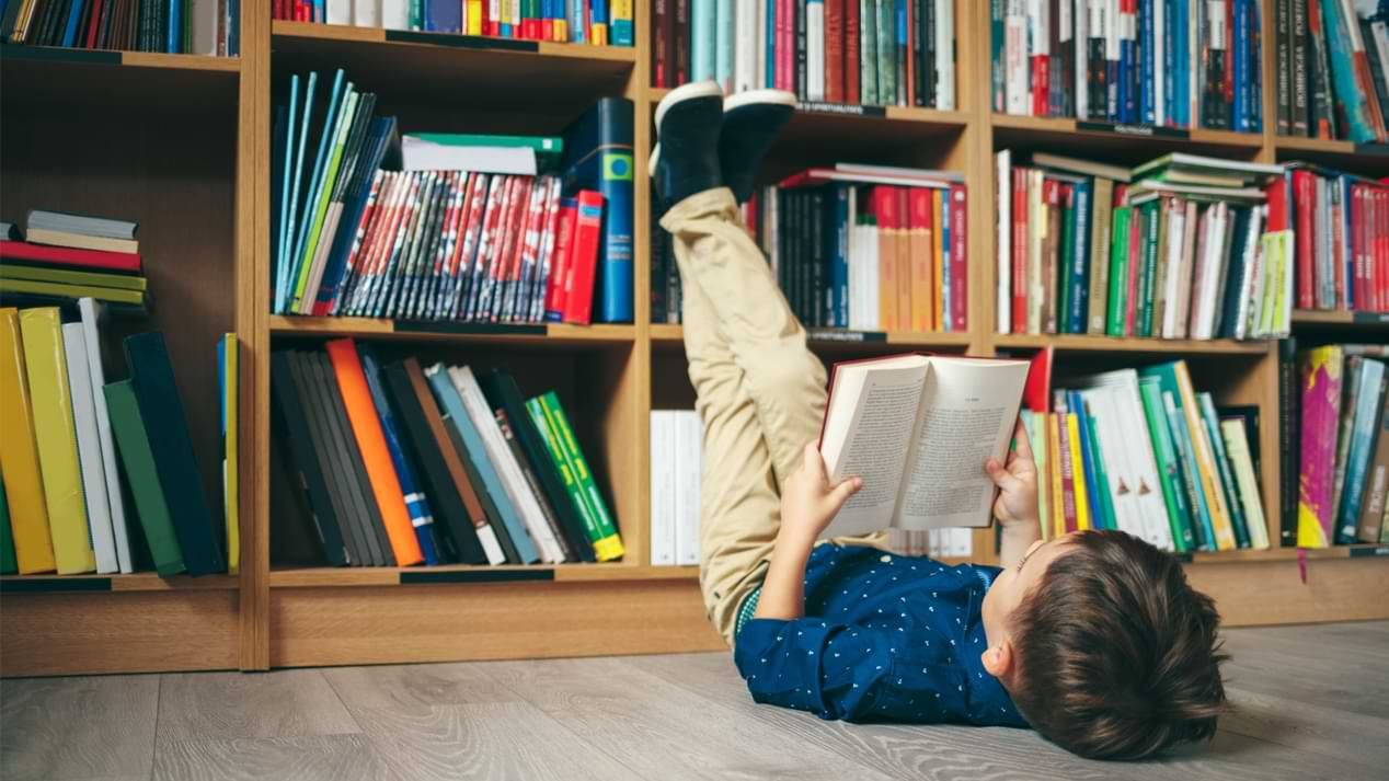 Ein Junge liegt vor einem vollen Bücherregal auf dem Rücken und liest ein Buch.