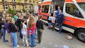 Vier Kinder und eine Frau stehen in der Tür eines Rettungswagens. Die Kinder halten ein Spendenschild hoch. Andere Kinder sehen ihnen zu.