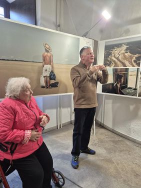 Führung durch die Ausstellung: Ein Museumsführer erklärt ein Bild der Ausstellung. Eine Frau sitzt auf dem Rollator. 