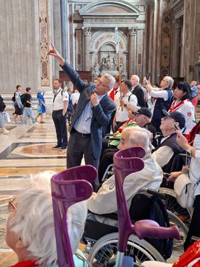 Eine Menschenmenge sieht nach oben, während ein Touristguide nach oben zeigt. Einige der Menschen sitzen im Rollstuhl oder haben Gehhilfen.
