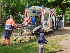 Ein Kind liegt auf einer Transportliege vor einem Rettungswagen. Andere Kinder schauen zu.