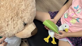 Ein Kind hält eine Stoffmöve mit "Papierschiene" in den Händen, beobachtet von einem Teddybären.