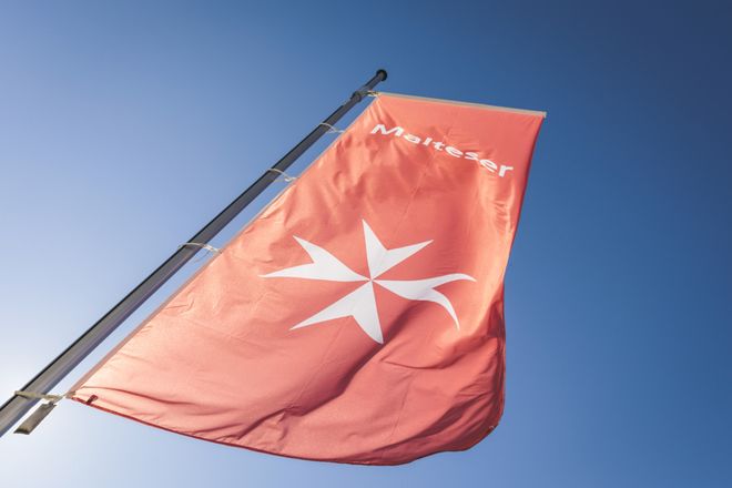 Rote Maltese-Flagge mit weißem Kreuzsymbol an einem Mast vor blauem Himmel.