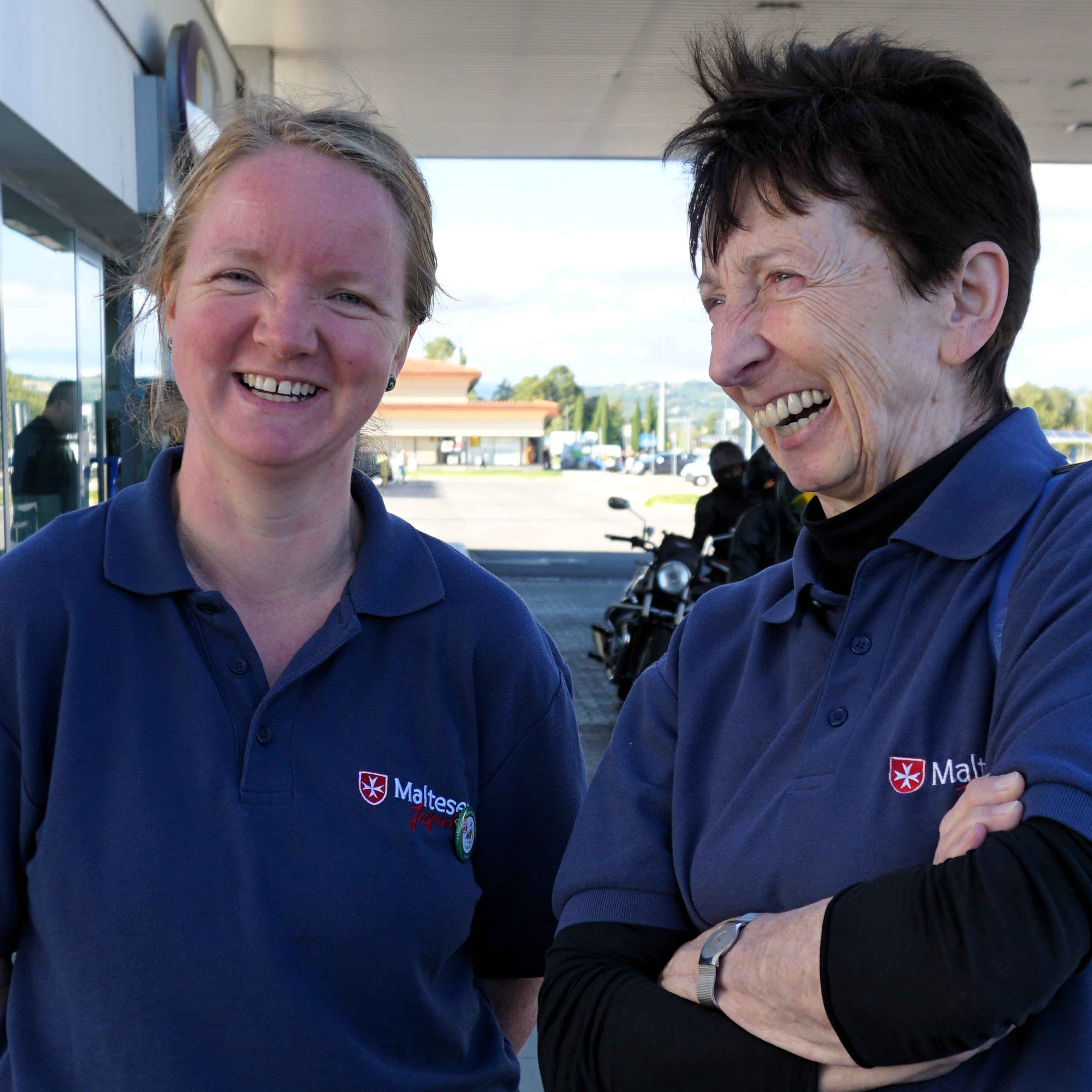 Zwei Personen in dunkelblauen Poloshirts mit Malteser Jugend Logo, eine mit verschränkten Armen, im Freien vor einem Gebäude.
