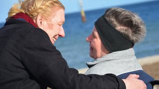 Eine Frau lächelt eine Frau im Rollstuhl an. Beide sind am Meer.