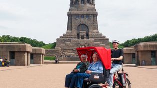 Eine Seniorin und ein Senior sitzen in der Rikscha. Ein Malteser Mitarbeitende sitzt auf dem Sattel. Im Hintergrund ist das Völkerschlachtdenkmal zu sehen.