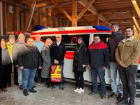 Eine Gruppe von Menschen steht in einer offenen Fahrzeughalle vor einem weiß-roten Einsatzfahrzeug mit blauen Signallichtern. Das Fahrzeug trägt die Aufschrift „Herzenswunsch-Krankenwagen“. Die Personen sind winterlich gekleidet und posieren gemeinsam für ein Gruppenfoto. Die Halle besteht aus Holzträgern und ist von natürlichem Licht erhellt.