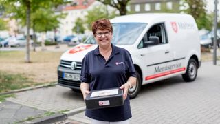 Mitarbeiterin des Malteser Menueservice trägt eine Warmhaltebox und lächelt in die Kamera.