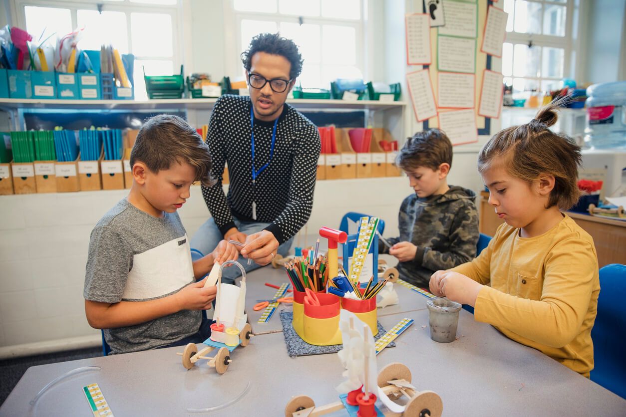 Junger Mann sitzt mit Schulkindern am Tisch und bastelt