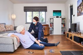 Eine ältere Dame sitzt in ihrer Wohnung auf dem Boden, angelehnt an das Sofa. Daneben ihre Brille und ein umgekipptes Glas. Sie trägt den Hausnotrufknopf als Kette um den Hals. Neben ihr kniet eine junge Dame von den Maltesern in Dienstbekleidung und kümmert sich um sie.