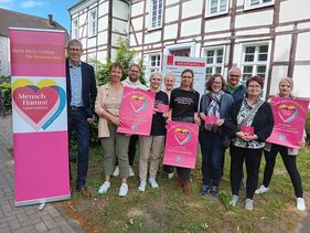 Eine Gruppe von zehn Menschen steht im Freien vor einem Fachwerkhaus mit weiß-brauner Fassade. Sie posieren für ein Gruppenfoto. Links im Bild steht ein großes, aufstellbares Banner mit bunten Herzformen und dem Text „Mensch Hamm! Lebe Freiheit! Mein Herz schlägt für Demokratie“. Einige Personen halten pinkfarbene Plakate oder Flyer mit ähnlichem Design in der Hand. Auf den Plakaten steht: „Wir sehen Dich und hören Dir zu!“ und „Wir sammeln DEINE Herzenswünsche für die Politik der Stadt Hamm“. Die Gruppe besteht aus Männern und Frauen verschiedenen Alters. Alle Personen wirken freundlich und lächeln in die Kamera. Die Stimmung ist positiv und gemeinschaftlich. Im Hintergrund ist ein weiteres Schild mit der Aufschrift „Geschäftsstelle“ zu sehen.