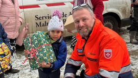Ein Mädchen hat ein weihnachtlich verpacktes Hilfspaket im Arm, daneben kniet Michael Soldanski, Leiter der Malteser Auslandshilfe