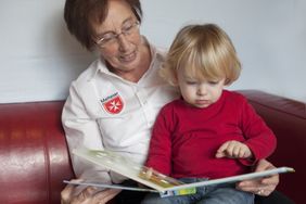 Eine ältere Frau hat ein Kleinkind auf dem Schoß. Beide schauen sich ein Bilderbuch an.