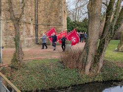 Vor dem historischen Kirchengebäude St. Johannes der Täufer gehen drei Bannerträger mit roten Bannern der Prozession voran.