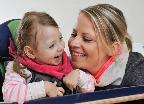 Eine lachende Frau schaut zu einem lachenden Kind, welches in einem Sitzt sitzt