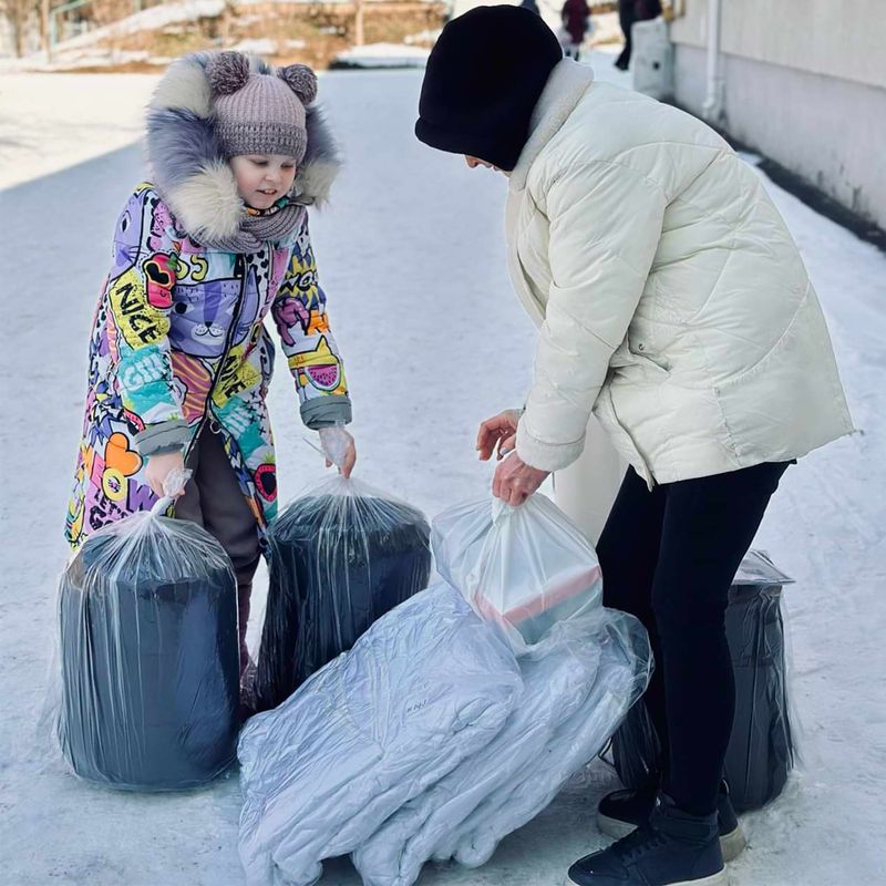 Eine Frau und ein Kind in Winterkleidung mit Säcken voller Textilien in einer winterlich-urbanen Umgebung.