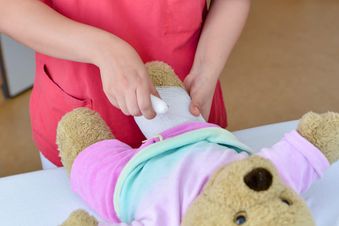 Pflegekraft in roter Kleidung wickelt ein Stofftier, demonstriert kindgerechte Versorgung in Krankenhausumgebung.