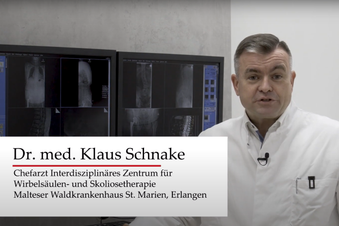 Arzt im weißen Kittel zeigt auf Monitore mit Wirbelsäulenbildern in einem Behandlungs-raum.