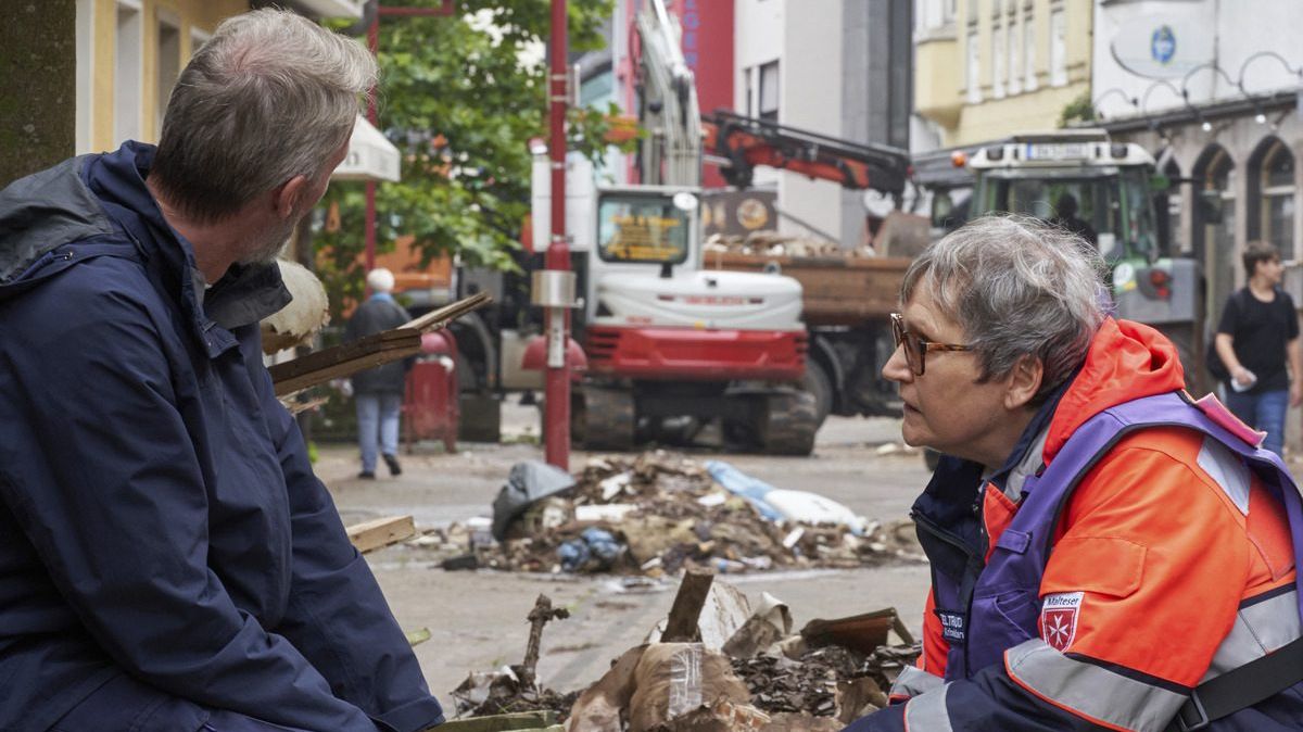 Eine Frau kniet vor einem Mann. Im Hintergrund Aufräumarbeiten.