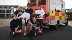 Sechs Jugendliche posieren in Malteser Kleidung und mit einem "abgerissenen Fuß" vor einem Rettungswagenu