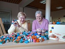 Zwei Personen sitzen an einem Tisch, der vollständig mit vielen bunt gestrickten Mini-Mützen bedeckt ist. Beide halten einige der gestrickten Mützen in den Händen. Rechts neben ihnen steht ein Karton mit dem Aufdruck „Mach Dich Mützlich“, verziert mit bunten Illustrationen und Strickmotiven. Die Szene wirkt freundlich und zeigt das gemeinsame Engagement für die Strickaktion.