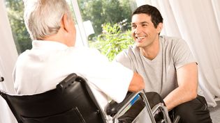 Junger Mann, der vor einer älteren Person im Rollstuhl kniet Malteser