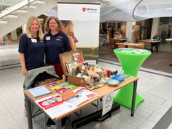 Zwei Malteser Frauen stehen hinter einem Stand und lächeln. Sie tragen dunkelblaue Malteserkleidung und informieren Besuchende.