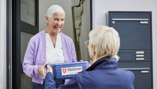 Ein älterer Herr sitzt an einem Tisch und lässt sich von einer Malteser-Mitarbeiterin ein Essens-Menü reichen.