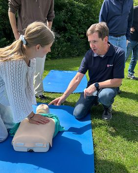 Zwei Personen und ein Erste-Hilfe-Dummy auf einer Matte. Die Eine Person zeigt der anderen, wie sie eine Herz-Druck-Massage richtig macht. Weitere Personen schauen zu.