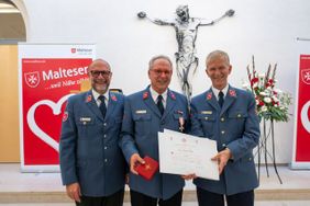 Drei Männer in blauen Uniformen stehen vor einem Altar, einer hält eine Urkunde und ein rotes Heft.