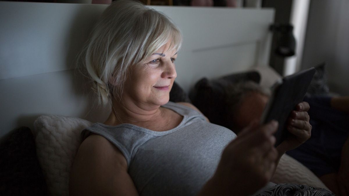 Ältere, lächelnde Frau liest im Bett mit einem E-Book-Reader.