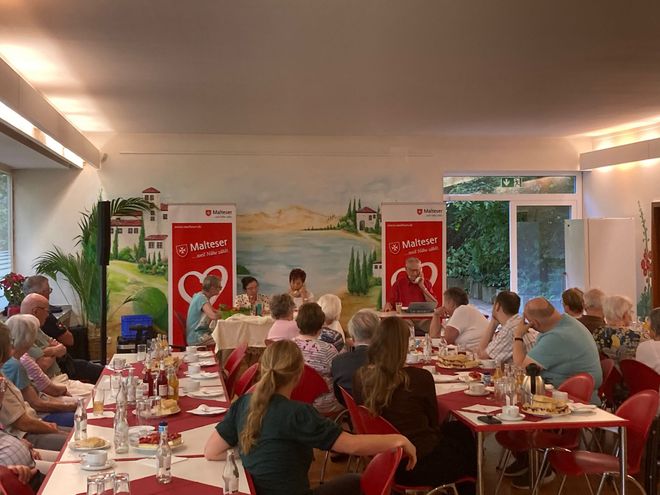 Zahlreiche Menschen sitzen an Tischen und sehen gespannt zu, wie die Referenten der Veranstaltung ein Telefongespräch nachstellen.