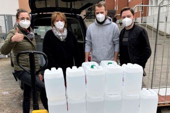 Vier Personen mit FFP2-Masken stehen hinter einer Palette mit Desinfektionsmittelkanistern vor Klinikgebäude und Fahrzeug.