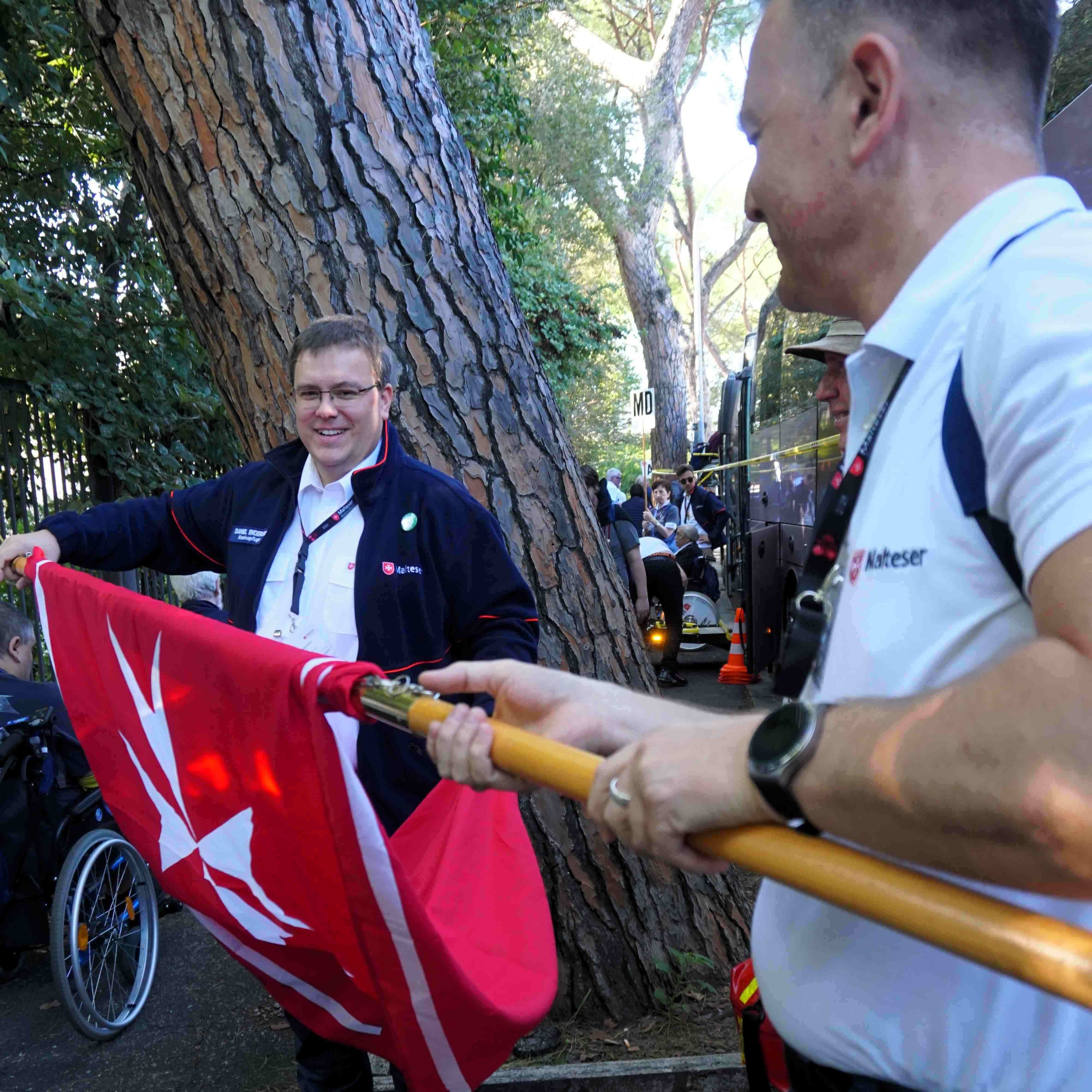Person hält rote Fahne mit weißem Symbol, im Hintergrund Baumstamm und weitere Personen im Freien.