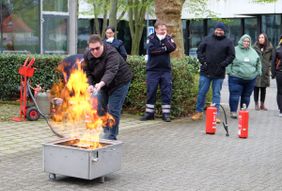 Zu sehen ist ein Feuer in einer für den Übungszweck vorgesehenen Wanne. Dahinter steht eine der Führungskräfte, d.h. ein junger Mann, der mittels eines Feuerlöschers den Löschvorgang beginnt. Im Hintergrund sind in sicherer Entfernung weitere zuschauende Führungskräfte zu sehen. 