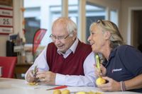 Ein älterer Mann und eine etwas jüngere Frau sitzen zusammen am Tisch und schälen Kartoffeln. Die Frau trägt ein blaues Malteser Shirt.