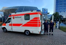 Zu sehen ist ein Rettungswagen mit Malteser-Logo. Rechts davon stehen zwei Männer in Malteser Dienstkleidung. Die Männer und der Rettungswagen stehen vor einer großen Halle.