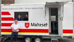 Anhänger der Mobilen Malteser Hilfer wo drei Helfer davor zu sehen sind