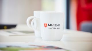 Weisse Malteser Tasse bedruckt mit Malteser Logo und Malteser–weil–Nähe–zählt-Schriftzug.