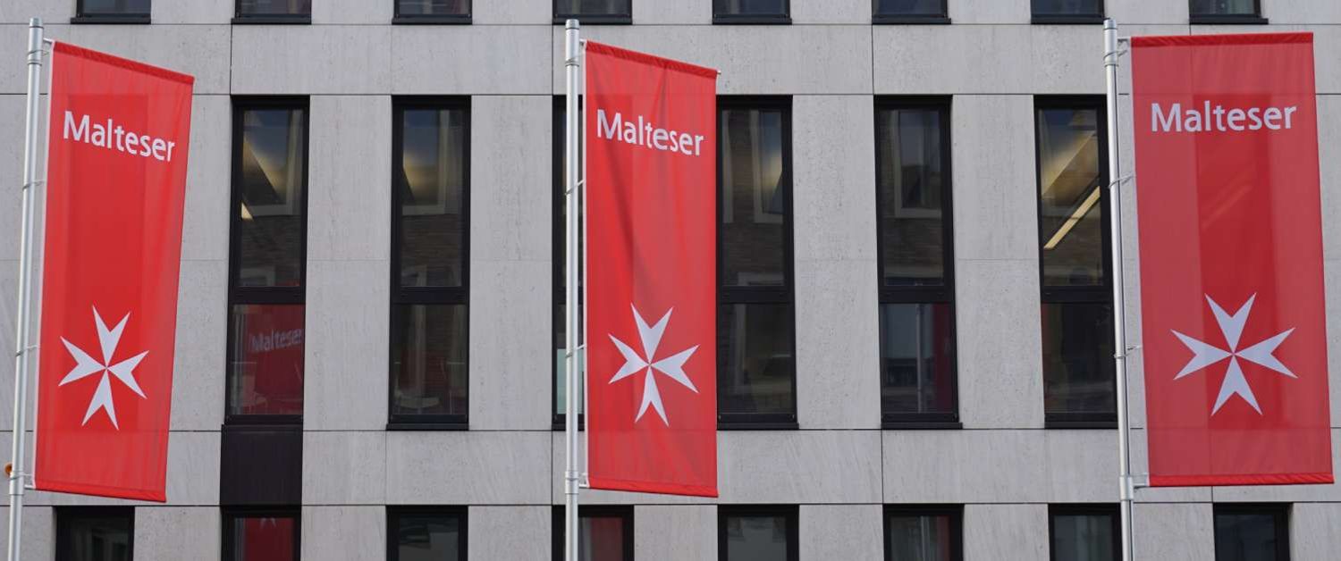 Drei rote Fahnen mit Malteser-Logo vor der Malteser-Zentrale