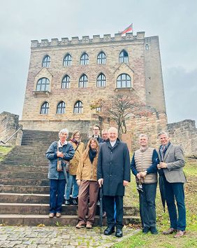 Eine Gruppe von sieben Personen steht vor einer mittelalterlichen Burg mit Treppe und Flagge.