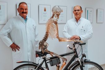 Zwei Ärzte in weißen Kitteln stehen neben einem Skelett auf einem Fahrrad.