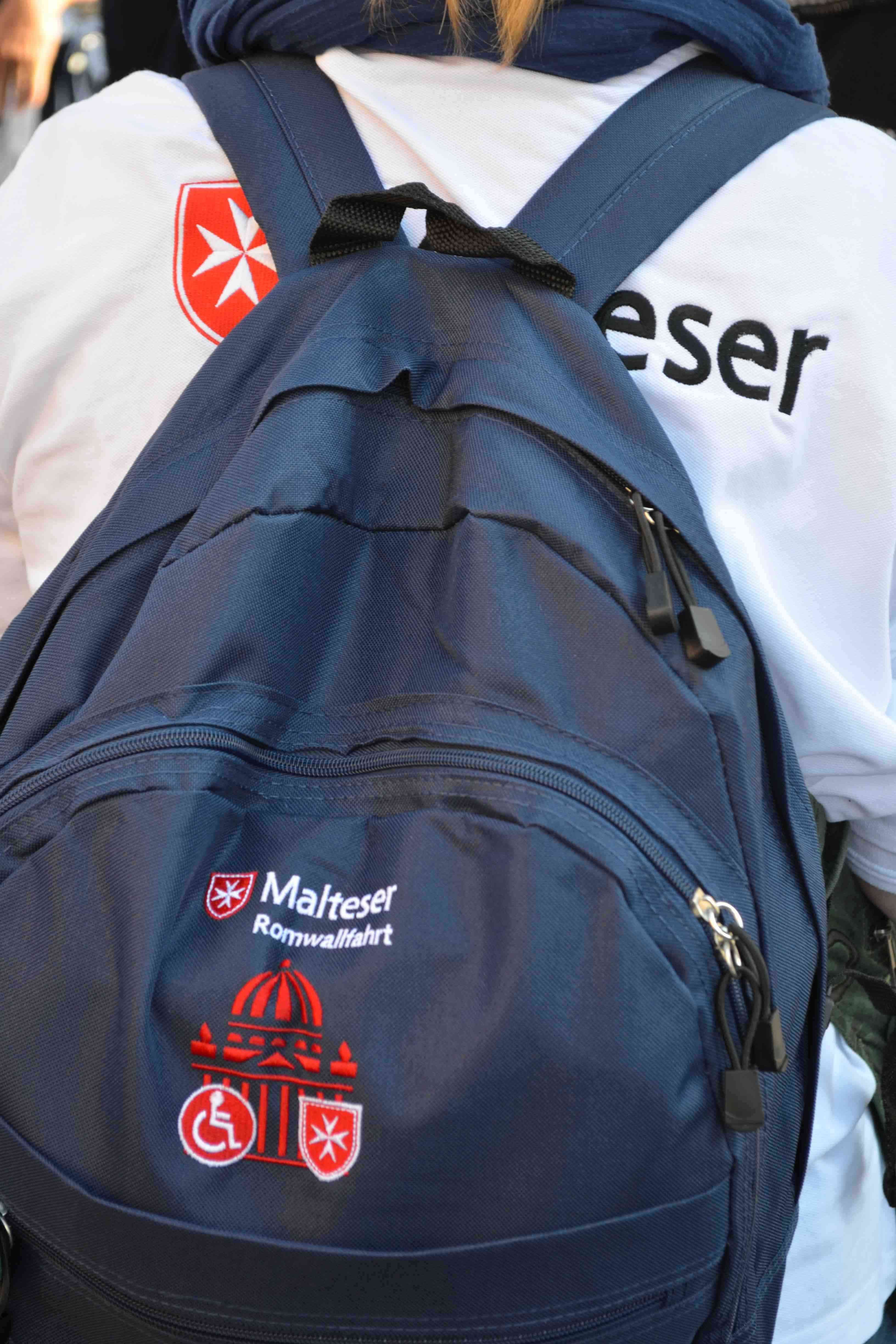 Blauer Rucksack mit mehreren Reißverschlusstaschen und roten Logos, getragen von einer Person in weißem Oberteil mit schwarzer Malteser Aufschrift.