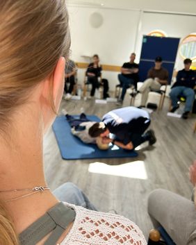 Seminarteilnehmerinnen und -teilnehmer schauen zu, wie der Referent in der Mitte des Raumes auf dem Boden an einer Erste-Hilfe-Puppe die Beatmung vorführt.