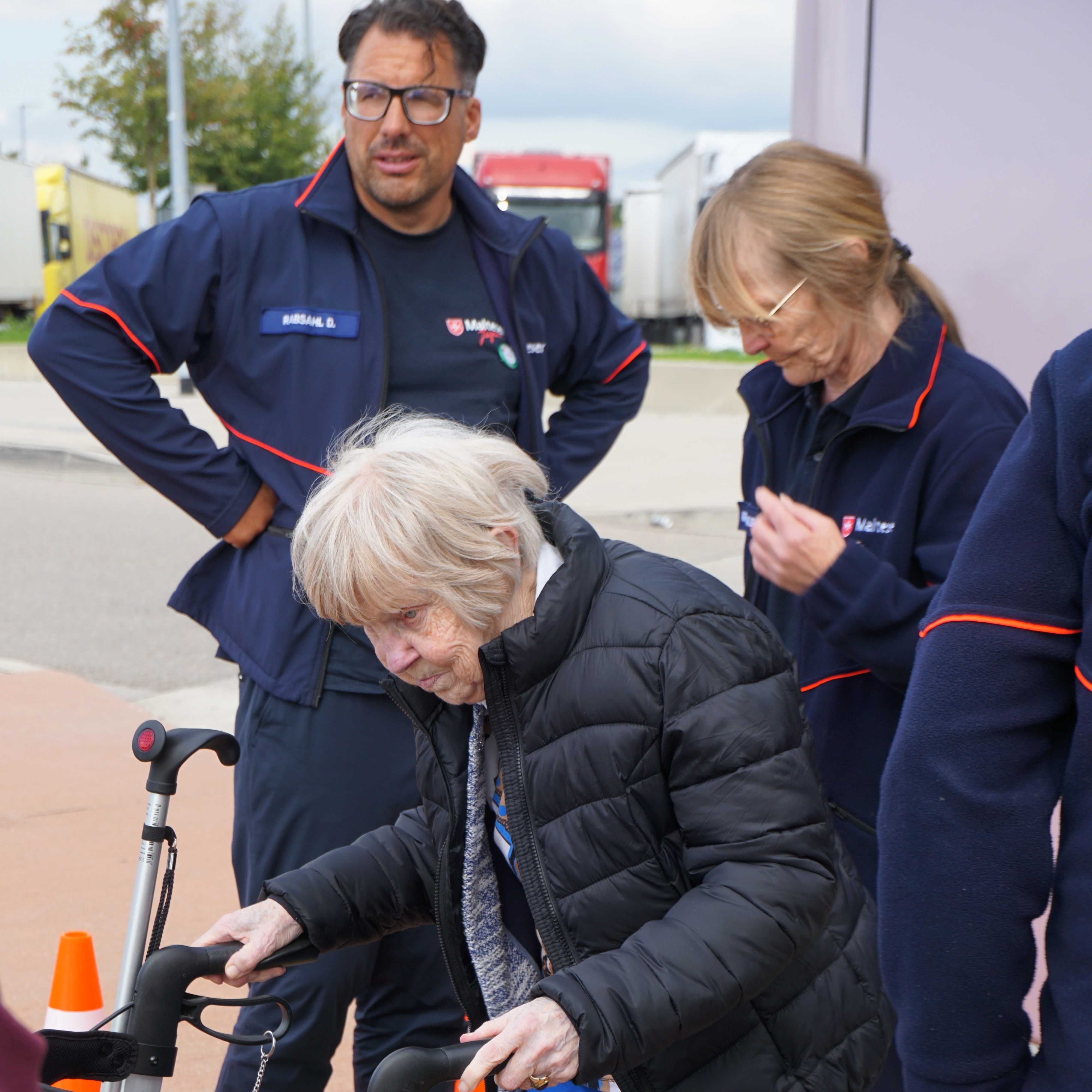 Ältere Person mit weißem Haar und schwarzer Jacke stützt sich auf einen Gehstock, umgeben von drei Personen in blauer Malteser Kleidung mit roten Akzenten.