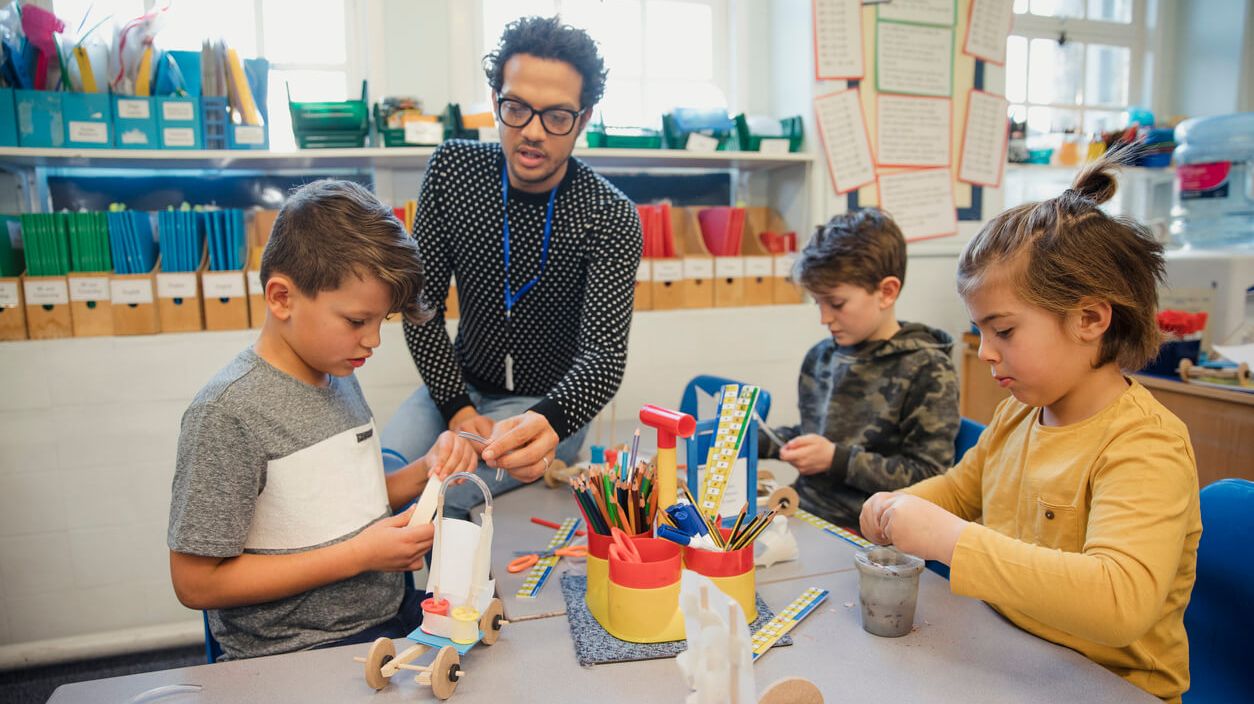 Junger Mann sitzt mit Schulkindern am Tisch und bastelt