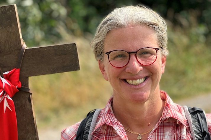 Portrait von Dörte Schrömges auf einer Wanderung mit einem Kreuz in der Hand und Wiese im Hintergrund