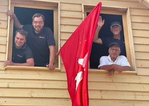 Vier Männer winken aus den Fenstern eines Holzhauses heraus. Am Haus ist eine Malteser Fahne befestigt.
