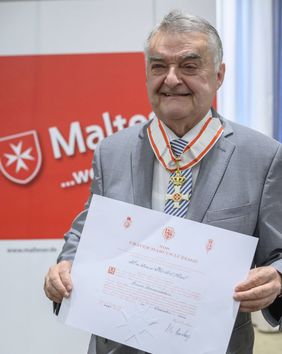 Herbert Reul trägt das Kommandeurkreuz und zeigt die Verleihungsurkunde der Regierung des Souveränen Malteser Ritterordens in Rom, die in lateinischer Sprache verfasst ist. 